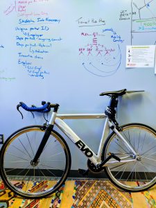 A bike inside a small office. Nonsensical writing on the whiteboard behind.