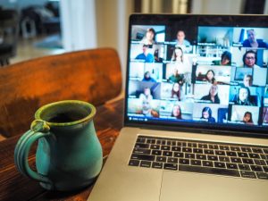 A laptop displaying a video conference page full of people, beside it a coffee mug.
